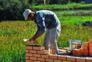 Construction worker laying bricks outdoors on a partially built wall with tools and materials beside him, set against a green, natural backdrop.