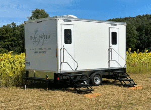 White 5 Unit Bonavita Luxury & Portable Lavatories restroom trailer parked in a sunny field beside blooming sunflowers, with dual entry stairs and trees in the background.
