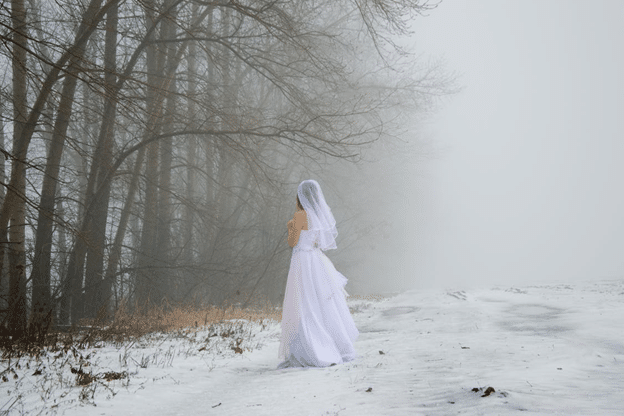Bride in white gown standing on a snowy path beside leafless winter trees.