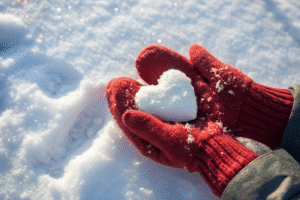 Red mittens holding a heart-shaped snowball on fresh snow.
