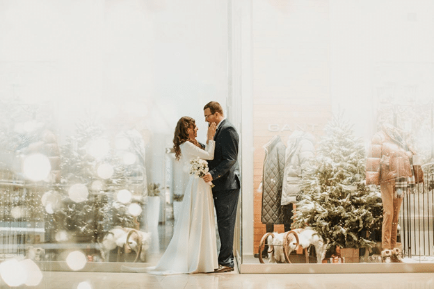 Bride and groom framed by a shop window with lit evergreens and winter coats.