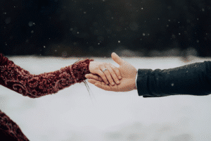 Close-up of two hands clasping in falling snow—red beaded sleeve and dark suit. Outdoor winter wedding.