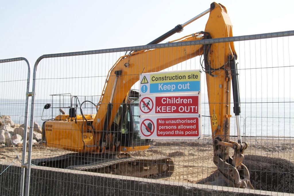 Temporary construction fencing with safety signs and an excavator behind it at a coastal jobsite.