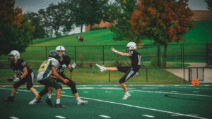 High school football punt on a turf field under fall trees.